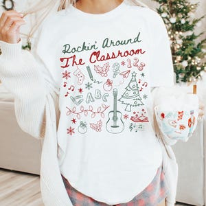 May include: White t-shirt with the text "Rockin' Around The Classroom" and a variety of Christmas-themed illustrations, including a Christmas tree, a guitar, a stocking, a candy cane, a pencil, and a book.