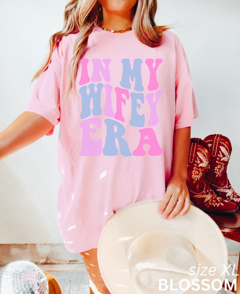 May include: Pink oversized t-shirt with the text "In My Wife Era" in a retro font with pink, blue, and purple colors. The shirt is being worn by a person and has a white hat and brown pants.