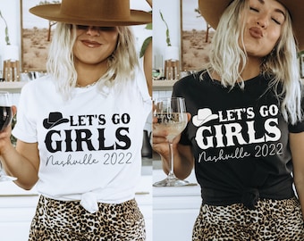 Può includere: Due donne indossano magliette bianche e nere con il testo "Let's Go Girls Nashville 2022" e una grafica di un cappello da cowboy. Le donne tengono bicchieri di vino e indossano cappelli da cowboy.