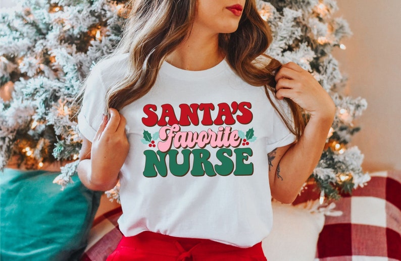 Puede incluir: Camiseta blanca con un gr&aacute;fico rojo, rosa y verde que dice "La enfermera favorita de Santa".