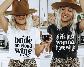 May include: Two women wearing hats and leopard print skirts. One woman is wearing a white t-shirt with the text "bride on cloud wine" in black. The other woman is wearing a black t-shirt with the text "girls just wanna have wine" in white. Both women are holding glasses of red wine.