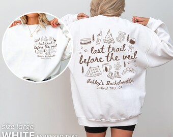 May include: White sweatshirt with brown text and camping-themed illustrations. The text reads "last trail before the veil" and "Ashley's Bachelorette, Joshua Tree, CA." The sweatshirt is size large.