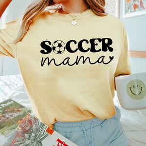 May include: A yellow t-shirt with the text "soccer mama" in black with a soccer ball graphic. The shirt is being worn by a person with blonde hair and blue shorts.