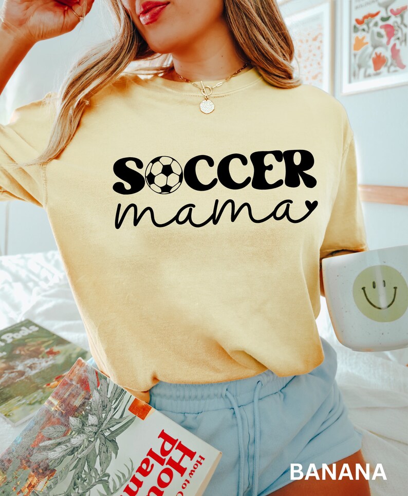 May include: A yellow t-shirt with the text "soccer mama" in black with a soccer ball graphic. The shirt is being worn by a person with blonde hair and blue shorts.