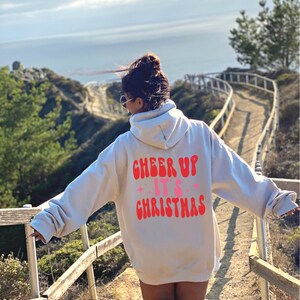 以下が含まれることがあります： 薄いグレーのフード付きスウェットシャツを着た人が、背中に「Cheer Up It's Christmas」という赤い文字とピンクの文字で書かれています。その人は、背景に海が見える木製の歩道の上に立っています。