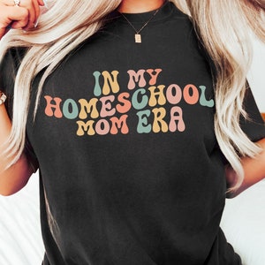 May include: Black t-shirt featuring the text "IN MY HOMESCHOOL MOM ERA" in a retro font. The text is in shades of pink, orange, blue, and yellow. The t-shirt is a plain black colour.