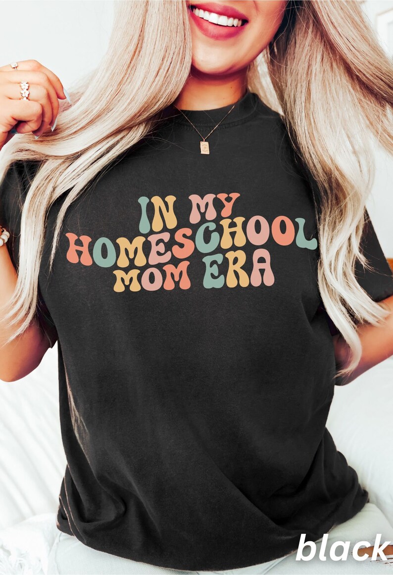 May include: Black t-shirt featuring the text "IN MY HOMESCHOOL MOM ERA" in a retro font. The text is in shades of pink, orange, blue, and yellow. The t-shirt is a plain black colour.