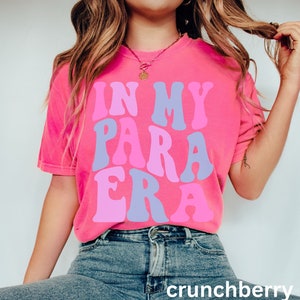 May include: A pink t-shirt with the text "IN MY PARA ERA" in a retro, colorful font.