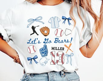 May include: White t-shirt with a baseball theme. It features a baseball player, a glove, baseballs, bats, bows, a jersey, a number "13", and the text "Let's Go Bears!" and "Miller".