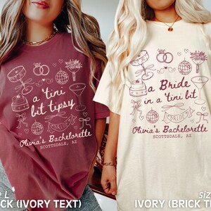Op de afbeelding: Twee T-shirts met een thema voor een vrijgezellenfeest. Het shirt aan de linkerkant is baksteenrood met ivoorwitte tekst die "a tini bit tipsy" en "Olivia's Bachelorette Scottsdale, AZ" zegt. Het shirt aan de rechterkant is ivoorwit met baksteenrode tekst die "a Bride in a tini bit" en "Olivia's Bachelorette Scottsdale, AZ" zegt.