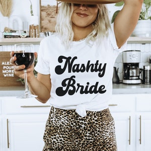Pu&ograve; includere: Una donna indossa una maglietta bianca con la scritta "Nasty Bride" in caratteri neri, un cappello fedora marrone e pantaloni a stampa leopardata e tiene un bicchiere di vino rosso.