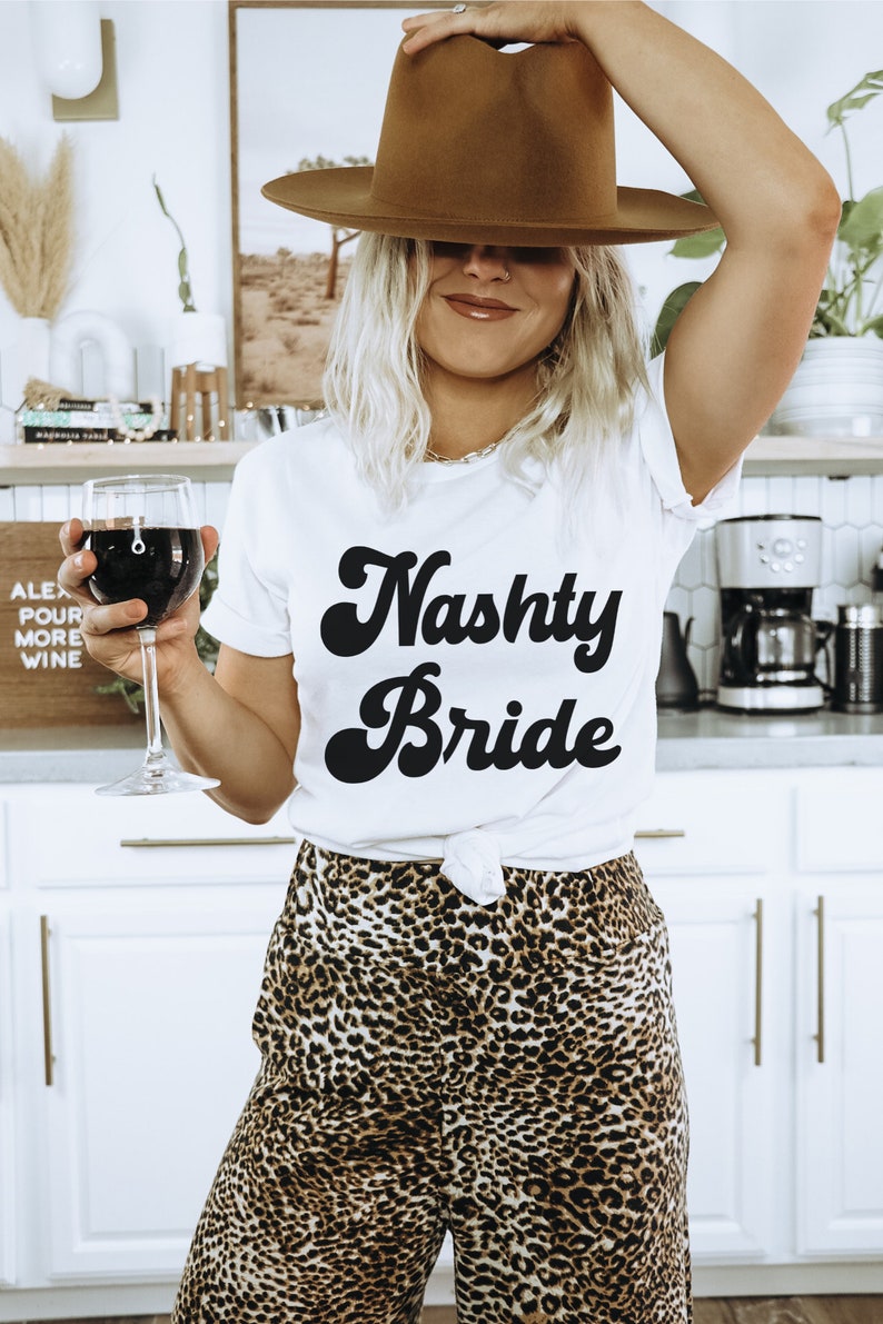 Pu&ograve; includere: Una donna indossa una maglietta bianca con la scritta "Nasty Bride" in caratteri neri, un cappello fedora marrone e pantaloni a stampa leopardata e tiene un bicchiere di vino rosso.
