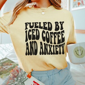 Peut inclure: T-shirt jaune clair avec l'inscription noire "FUELED BY ICED COFFEE AND ANXIETY". La personne tient une tasse blanche avec un smiley et un livre. Le t-shirt est de couleur jaune clair.