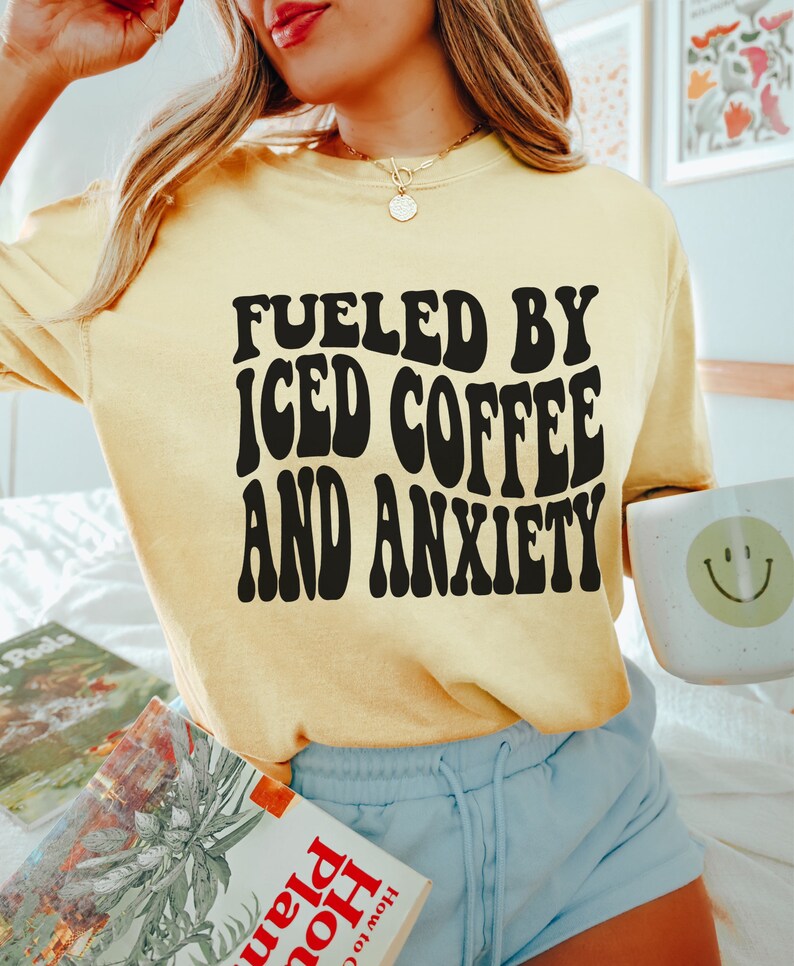 Peut inclure: T-shirt jaune clair avec l'inscription noire "FUELED BY ICED COFFEE AND ANXIETY". La personne tient une tasse blanche avec un smiley et un livre. Le t-shirt est de couleur jaune clair.