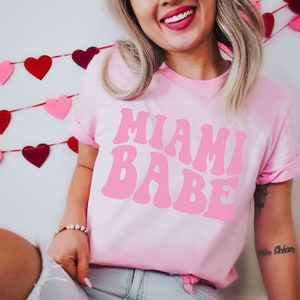Pode incluir: Uma camiseta rosa com o texto "Miami Babe" em uma fonte rosa retr&ocirc;. A camiseta est&aacute; sendo usada por uma pessoa com cabelo loiro comprido. Uma guirlanda de cora&ccedil;&otilde;es vermelhos e rosa est&aacute; no fundo.