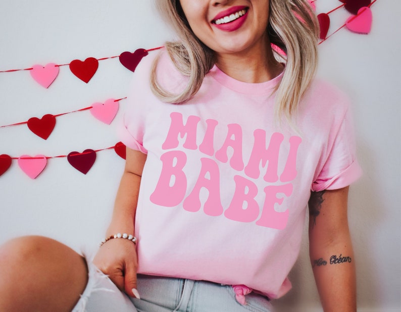 Pode incluir: Uma camiseta rosa com o texto "Miami Babe" em uma fonte rosa retr&ocirc;. A camiseta est&aacute; sendo usada por uma pessoa com cabelo loiro comprido. Uma guirlanda de cora&ccedil;&otilde;es vermelhos e rosa est&aacute; no fundo.