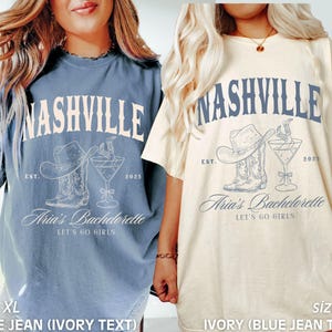 May include: Two oversized t-shirts, one blue and one ivory, each featuring a Nashville-themed design. The design includes the word "Nashville," a cowboy hat, boots, and a cocktail glass. The text "Aria's Bachelorette" and "Let's Go Girls" are also present.