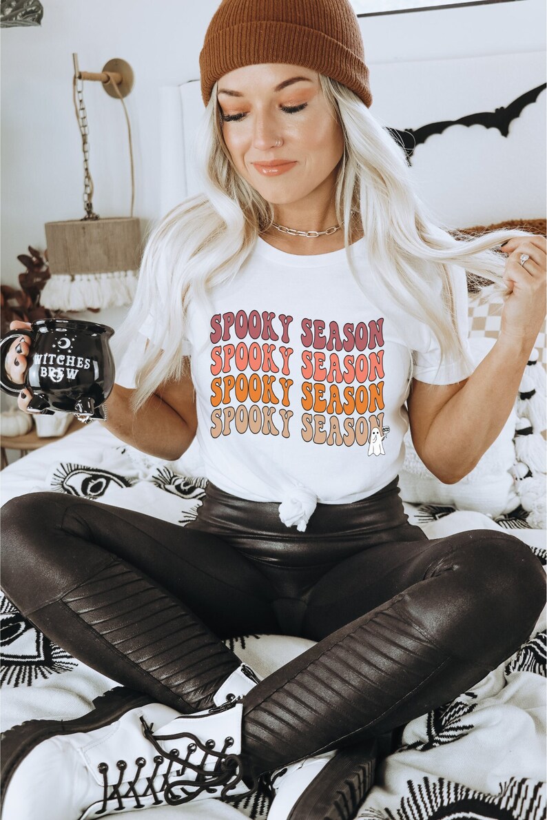 May include: White t-shirt with the text "Spooky Season" in a retro font, layered in shades of pink, orange, and beige. The shirt features a small ghost graphic. The person is wearing a brown beanie, black leather trousers, and white boots.
