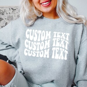 Pu&ograve; includere: Una felpa grigio chiaro con la scritta "CUSTOM TEXT" in bianco e con un design ondulato, ripetuta tre volte. La felpa casual &egrave; adatta all'uso quotidiano. L'immagine mostra un primo piano della parte superiore del corpo.