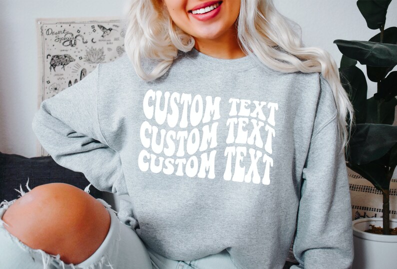 Pu&ograve; includere: Una felpa grigio chiaro con la scritta "CUSTOM TEXT" in bianco e con un design ondulato, ripetuta tre volte. La felpa casual &egrave; adatta all'uso quotidiano. L'immagine mostra un primo piano della parte superiore del corpo.