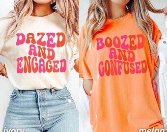 May include: Two t-shirts with retro-style text. The first shirt is ivory with the text "Dazed and Engaged" in pink and orange. The second shirt is orange with the text "Boozed and Confused" in pink and red.