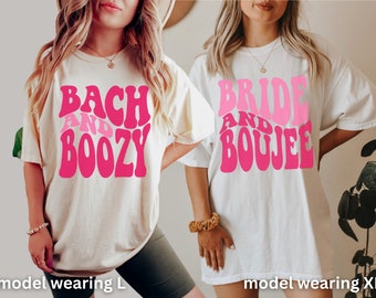 May include: Two white t-shirts with pink text. The left shirt says "Bach and Boozy" and the right shirt says "Bride and Boujee". The model on the left is wearing a size L and the model on the right is wearing a size XL.
