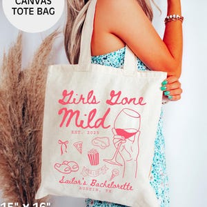 May include: A natural canvas tote bag with the text "Girls Gone Mild EST. 2025" in pink lettering. The bag also features a pink illustration of a hand holding a glass of wine, along with other pink illustrations of a pizza slice, popcorn, a pair of flip flops, and an ice cream cone. The text "Sailor's Bachelorette Austin, TX" is also printed in pink lettering.