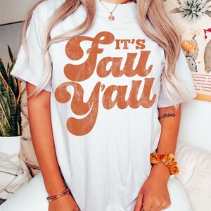 May include: White t-shirt with a brown graphic that says "It's Fall Y'all" in a retro font.