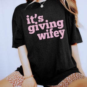 May include: Black oversized t-shirt with pink text that reads "it's giving wifey".