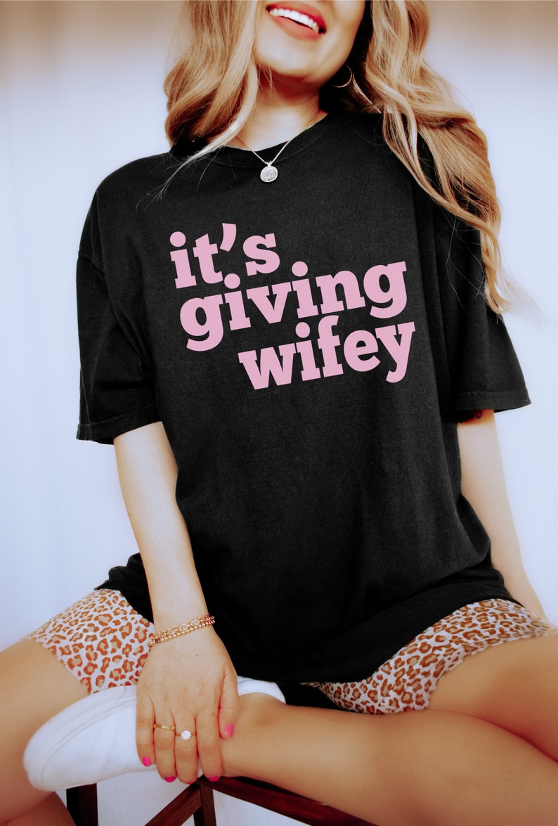 May include: Black oversized t-shirt with pink text that reads "it's giving wifey".