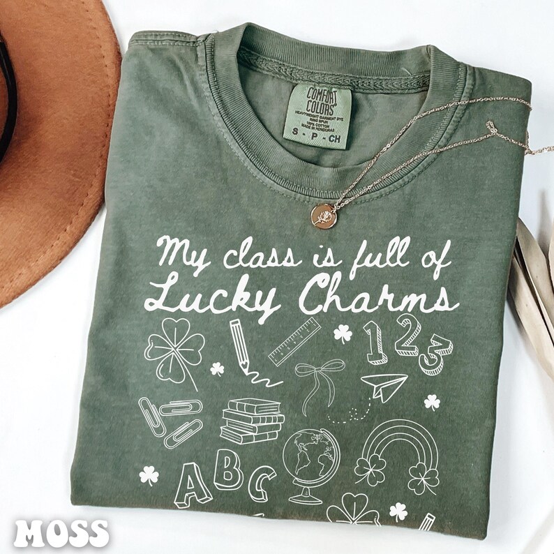 May include: Green t-shirt with the text "My class is full of Lucky Charms" in white. The shirt has a variety of school-themed illustrations, including a pencil, ruler, globe, books, paperclip, shamrocks, and the letters ABC and numbers 123.
