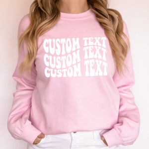 Pu&ograve; includere: Una felpa rosa chiaro a maniche lunghe. La parte anteriore della felpa presenta le parole "CUSTOM TEXT" in un carattere bianco ondulato, ripetute tre volte verticalmente. La felpa ha un girocollo e polsini a coste.
