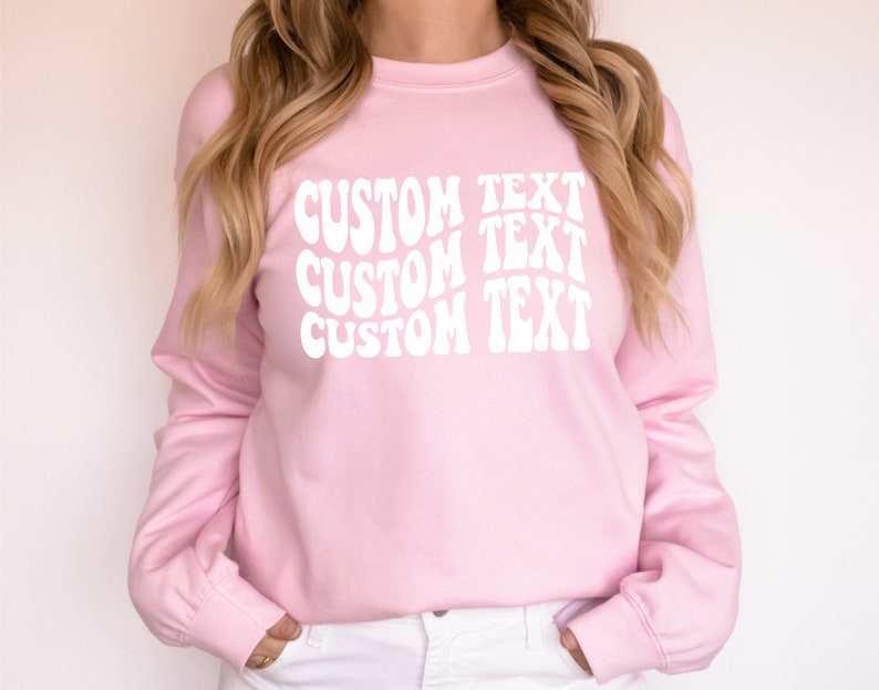 Pu&ograve; includere: Una felpa rosa chiaro a maniche lunghe. La parte anteriore della felpa presenta le parole "CUSTOM TEXT" in un carattere bianco ondulato, ripetute tre volte verticalmente. La felpa ha un girocollo e polsini a coste.