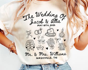 May include: Ivory t-shirt with black text and illustrations. The text reads "The Wedding of Jacob & Allie May 18th, 2025 Mr. & Mrs. Williams Nashville, TN". The illustrations include wedding rings, a cake, a church, a car, doves, champagne glasses, a bouquet of flowers, and a disco ball.
