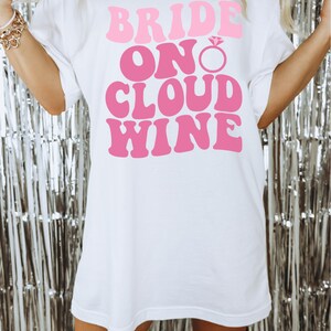 Peut inclure: T-shirt blanc avec le texte "Bride On Cloud Wine" en lettres roses avec un graphique de bague de fian&ccedil;ailles en diamant rose.