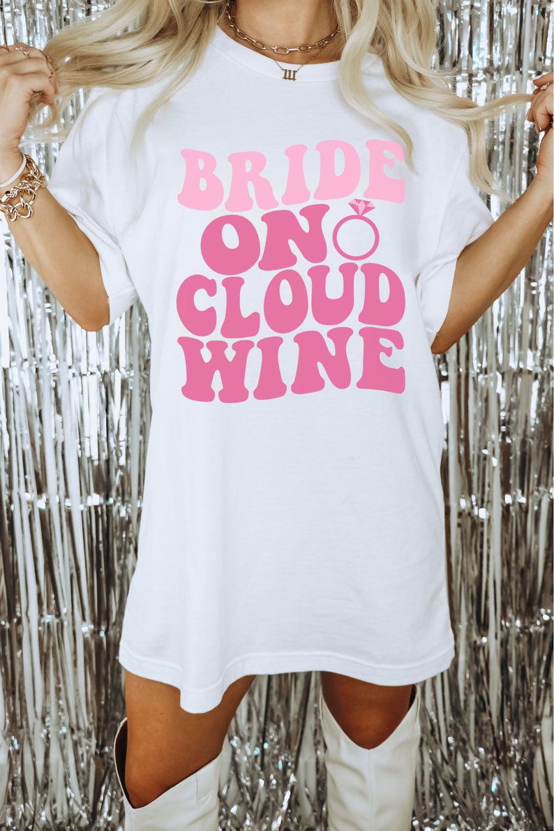 Peut inclure: T-shirt blanc avec le texte "Bride On Cloud Wine" en lettres roses avec un graphique de bague de fian&ccedil;ailles en diamant rose.