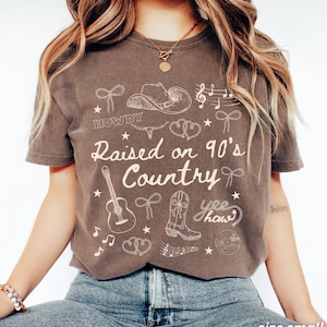 May include: Espresso-colored t-shirt with the text "Raised on 90's Country" and country-themed graphics. The design includes a cowboy hat, guitar, boots, musical notes, and the word "Howdy". The shirt is size small.