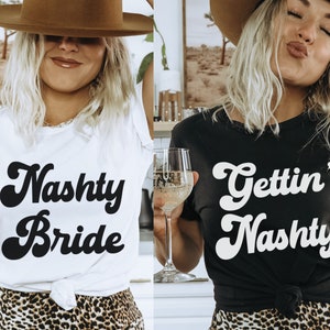 Pu&ograve; includere: Due donne indossano magliette bianche e nere con il testo "Naughty Bride" e "Gettin' Naughty" in un font retr&ograve;. Entrambe le donne indossano cappelli da cowboy marroni e gonne a stampa leopardata.
