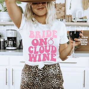 Peut inclure: Un t-shirt blanc avec le texte "Bride On Cloud Wine" en rose r&eacute;tro. Le t-shirt pr&eacute;sente un graphique de bague de fian&ccedil;ailles en diamant rose. La personne portant le t-shirt tient un verre de vin rouge.