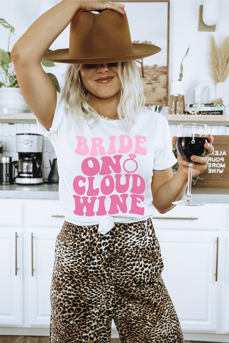 Peut inclure: Un t-shirt blanc avec le texte "Bride On Cloud Wine" en rose r&eacute;tro. Le t-shirt pr&eacute;sente un graphique de bague de fian&ccedil;ailles en diamant rose. La personne portant le t-shirt tient un verre de vin rouge.