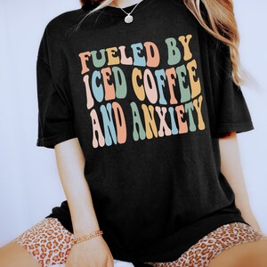 Peut inclure: T-shirt noir avec l'inscription "FUELED BY ICED COFFEE AND ANXIETY" dans un style r&eacute;tro color&eacute;. Le t-shirt a une coupe ample, parfaite pour un look d&eacute;contract&eacute;. L'ensemble d&eacute;gage une ambiance vintage et amusante.