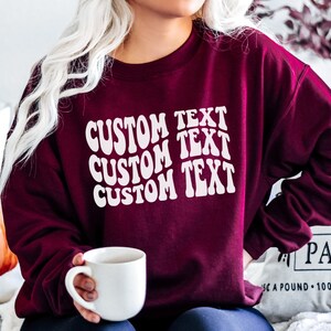 Pu&ograve; includere: Felpa bordeaux con la scritta "CUSTOM TEXT" in bianco, con un font ondulato. La persona tiene in mano una tazza bianca. Lo sfondo include una zucca e un cuscino.