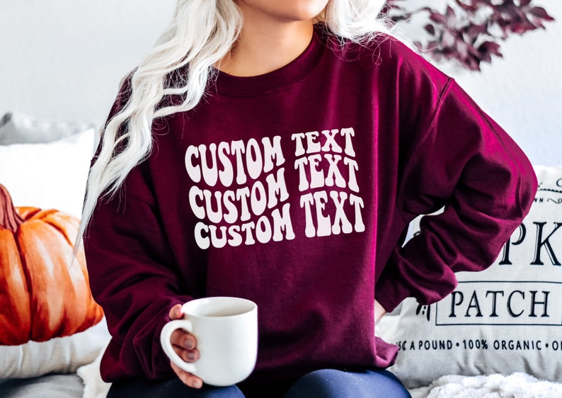 Pu&ograve; includere: Felpa bordeaux con la scritta "CUSTOM TEXT" in bianco, con un font ondulato. La persona tiene in mano una tazza bianca. Lo sfondo include una zucca e un cuscino.