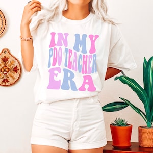 Peut inclure: T-shirt blanc avec le texte "In My P.D. Teacher Era" en police r&eacute;tro rose et bleue.
