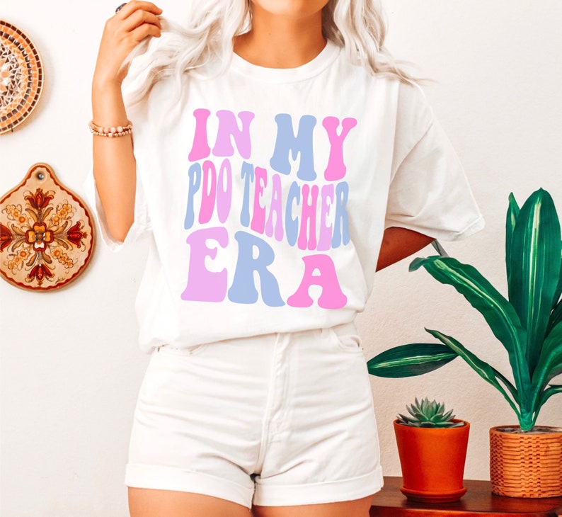 Peut inclure: T-shirt blanc avec le texte "In My P.D. Teacher Era" en police r&eacute;tro rose et bleue.