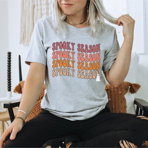 May include: A light grey t-shirt featuring the text "Spooky Season" in a retro font, stacked in pink, orange, and peach. A small ghost graphic is on the bottom right. The shirt is part of a Halloween-themed collection.