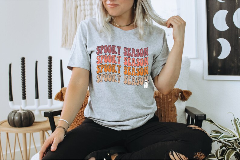 May include: A light grey t-shirt featuring the text "Spooky Season" in a retro font, stacked in pink, orange, and peach. A small ghost graphic is on the bottom right. The shirt is part of a Halloween-themed collection.