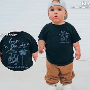 Op de afbeelding: Zwart T-shirt met de tekst "One in the Sun" en "Elio's Pool Party" in gewassen denim. Het shirt heeft een strandsc&egrave;ne-afbeelding met een parasol, stoel en palmboom. De achterkant van het shirt heeft ook hetzelfde ontwerp.
