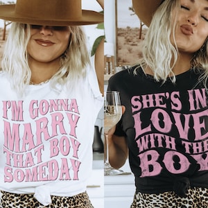 Puede incluir: Dos mujeres con camisetas con texto rosa. La camiseta blanca dice "Voy a casarme con ese chico alg&uacute;n d&iacute;a". La camiseta negra dice "Ella est&aacute; enamorada del chico".