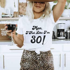 May include: A white t-shirt with the text "Man, I feel like I'm 30!" in a retro font. The person wearing the shirt is holding a glass of red wine and wearing a brown fedora and leopard print pants.